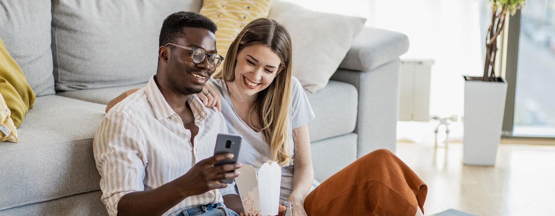 a man and woman with take out, sit against a couch on their living room floor and watch at their cell phone