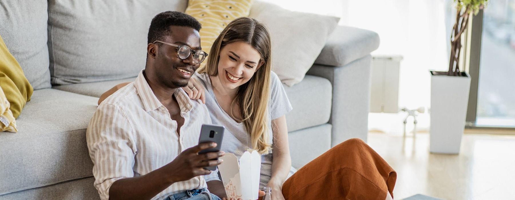 a man and woman with take out, sit against a couch on their living room floor and watch at their cell phone